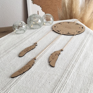 May include: A wooden dreamcatcher with three wooden feathers hanging from it. The feathers are decorated with the words "6.620 cm", "4.7 cm", and "6.4 cm".
