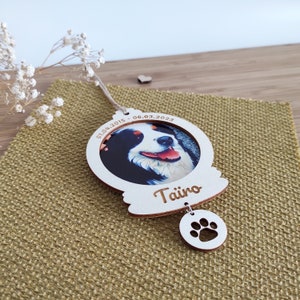 Può includere: Ciondolo in legno bianco con una foto di un cane all'interno di un cerchio. Il ciondolo è inciso con le date "01.04.2015 - 06.03.2023" e il nome "Taino". Un piccolo ciondolo in legno a forma di impronta di zampa pende dal ciondolo.