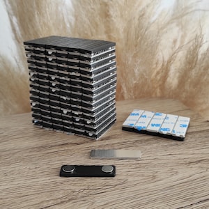 May include: A stack of black magnetic squares, a silver metal magnetic strip, and a black magnetic strip with a silver metal accent. The magnetic squares are stacked on a wooden surface. The magnetic strips are on the wooden surface.