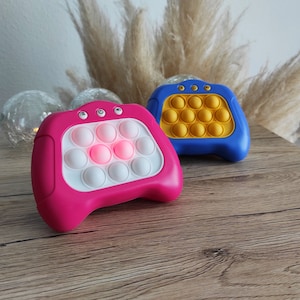 May include: Two pop-it fidget toys, one pink and one blue, with a light-up centre on the pink toy. Both toys have a textured surface and are shaped like a controller.
