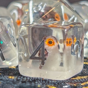 May include: Set of six clear resin dice with embedded owl and leaf designs. The dice have silver numbering and are displayed on a dark, textured surface. The dice are a unique set for tabletop gaming.