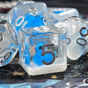 May include: Set of clear dice with blue duck-shaped inclusions and black numbering. The dice are in various polyhedral shapes, including a D20, D6, and D10. The dice are set against a dark background with a hint of blue and gold.