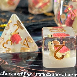 Pode incluir: Um conjunto de dados de resina transparente com numeração dourada e elementos decorativos. Alguns dados apresentam um design de tartaruga vermelha e dourada, outros detalhes florais vermelhos. Dados para jogos de tabuleiro.