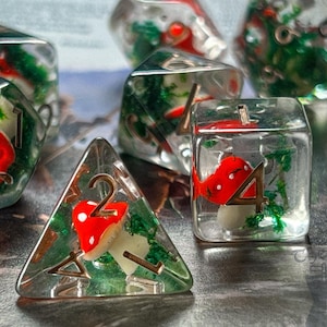 Può includere: Set di sette dadi in resina trasparente con inclusioni di funghi rossi e bianchi e fogliame verde. I dadi sono di varie forme, tra cui un d20, d12, d10, d8, d6, d4 e un cubo. I numeri sono dipinti in oro.