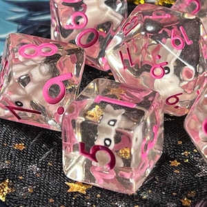 May include: A set of seven clear, pink-numbered dice with a unicorn figurine inside each die. The dice are in various shapes, including a d20, d12, d10, d8, d6, and d4. The numbers are in a bright pink color.