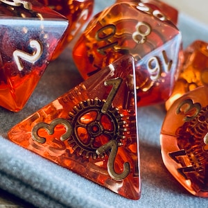 May include: A set of six orange and gold dice with gears and numbers on the faces. The dice are made of clear plastic and have a metallic gold finish.