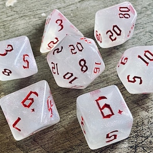 May include: Set of seven white dice with red numbers. The dice are translucent and have a glittery finish. The numbers on the dice are in a gothic font.