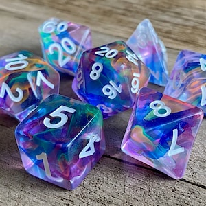 May include: A collection of seven polyhedral dice with a colorful, swirling design. The dice are transparent with shades of blue, purple, green, and orange. Each die has white numbers. These dice are designed for tabletop gaming.