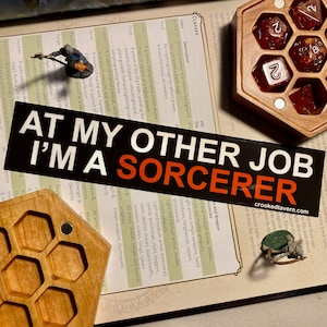 May include: A close-up of a tabletop role-playing game (RPG) setup with a miniature figure of a sorcerer, dice, and a sticker that reads "At my other job I'm a sorcerer". The sticker is placed on a book with a list of character classes and their descriptions.