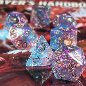 Op de afbeelding: Een set van zeven iriserende dobbelstenen voor tabletop games. De dobbelstenen zijn transparant met blauwe en paarse glitter en gouden cijfers. De dobbelstenen bevatten een D4, D6, D8, D10, D12, D20 en een percentieldobbelsteen.