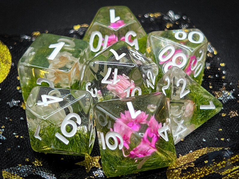 May include: A set of six clear resin dice with white numbers and pink and green pressed flowers inside. The dice are shaped like a traditional cube and have numbers 1 through 6 on each side.
