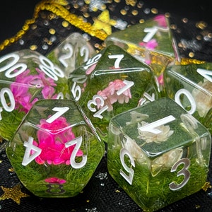 May include: Six clear resin dice with white numbers and pressed flowers and greenery inside. The dice are on a black background with gold glitter.