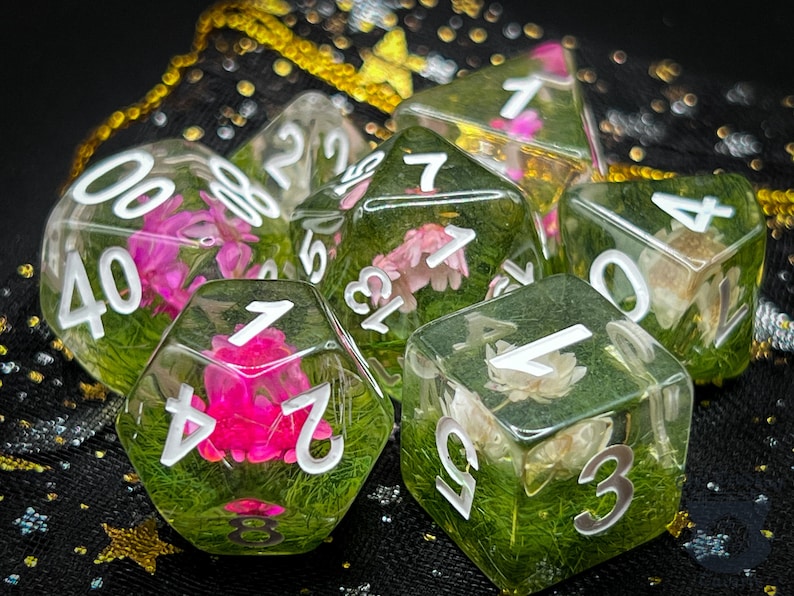 May include: Six clear resin dice with white numbers and pressed flowers and greenery inside. The dice are on a black background with gold glitter.