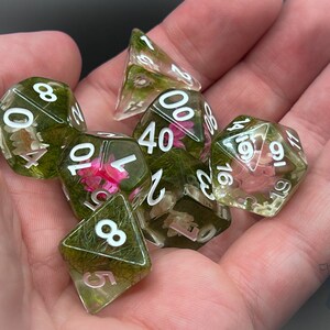 May include: A set of six green and clear dice with white numbers and embedded flowers and greenery. The dice are in a hand.