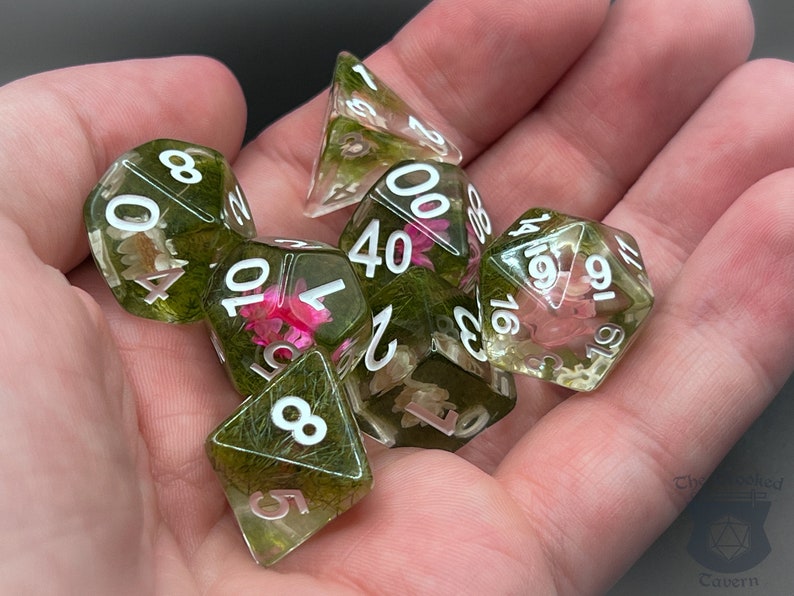 May include: A set of six green and clear dice with white numbers and embedded flowers and greenery. The dice are in a hand.
