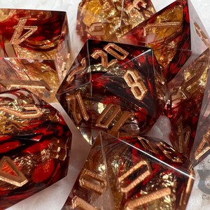 Pu&ograve; includere: Set di sette dadi poliedrici. Ogni dado &egrave; trasparente con vortici rossi e neri, accenti in lamina d'oro e numeri color bronzo. I dadi sono progettati per giochi di ruolo da tavolo.