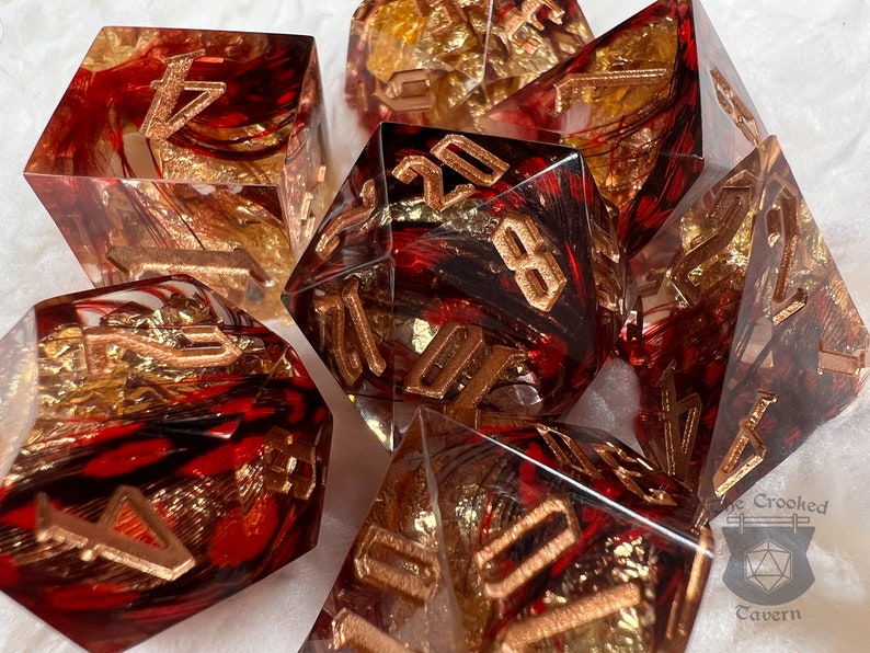 Pu&ograve; includere: Set di sette dadi poliedrici. Ogni dado &egrave; trasparente con vortici rossi e neri, accenti in lamina d'oro e numeri color bronzo. I dadi sono progettati per giochi di ruolo da tavolo.