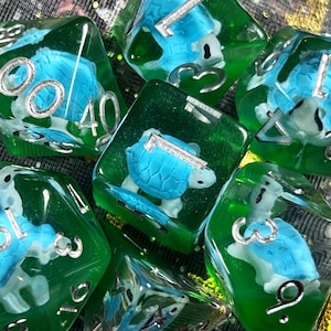 May include: Set of seven polyhedral dice with a green and clear resin. Each die features a blue turtle figurine and silver numbering. The dice are designed for tabletop role-playing games.