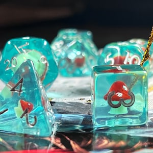 May include: A set of clear, teal-colored dice with red mushroom inclusions. The dice are of various shapes, including a D20, a D6, and a D4. The numbers are in a dark color, and the mushrooms have white spots.