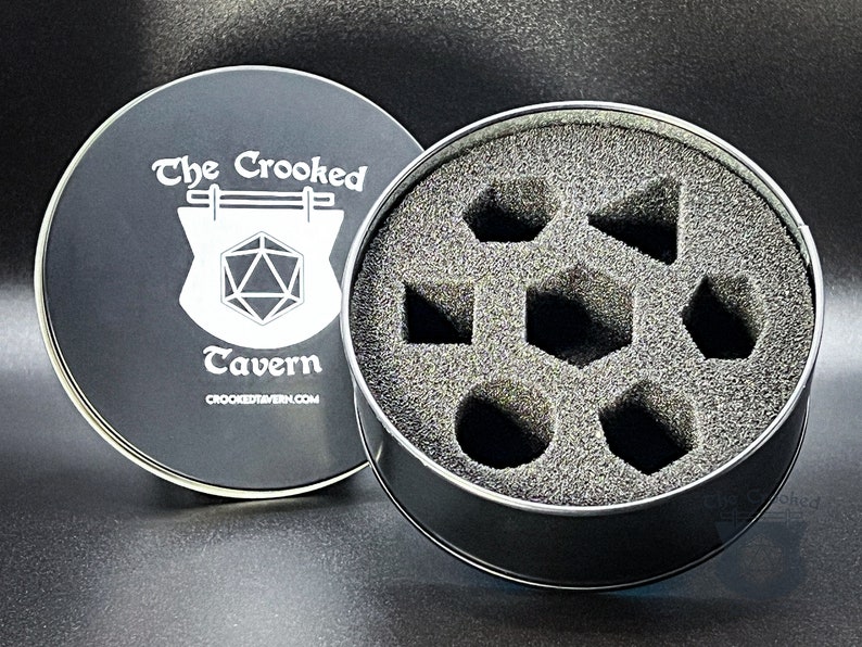 May include: A black metal tin with a foam interior for storing dice. The lid features a white logo that says "The Crooked Tavern" with a stylized image of a tavern sign and a 20-sided die.