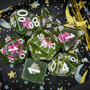 May include: A set of six clear dice with green and white floral accents. The dice are on a black background with gold stars and moons.
