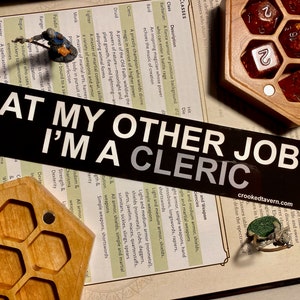 May include: A black and white sticker with the text "At my other job I'm a cleric" on a table with a Dungeons and Dragons rulebook and dice.