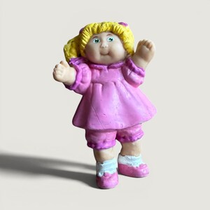 May include: A small figurine of a doll with yellow hair styled in pigtails, wearing a pink dress and matching pink shorts. The doll has green eyes, a smiling expression, and is wearing white socks with pink shoes, with both arms raised upward.
