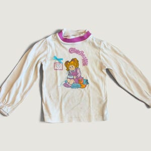 Op de afbeelding: Een wit shirt met lange mouwen en een paarse opstaande kraag. De voorkant heeft een afbeelding van een Cabbage Patch Kids-figuur met oranje haar, een kleine oranje kat en een blauw strikje. De tekst Cabbage Patch Kids is in het roze gedrukt.