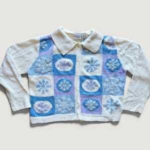 May include: A cream-coloured button-down cardigan featuring a patchwork design of blue, light blue, and lavender squares. Each square contains an embroidered snowflake or circular pattern. The garment has a ribbed collar and long sleeves.