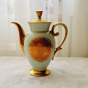 Puede incluir: Una tetera de porcelana blanca con detalles dorados y un diseño floral detallado. La tetera tiene una tapa y un asa.