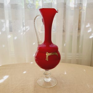 Könnte beinhalten: Eine leuchtend rote Glaskaraffe mit einem Griff und einem Sockel aus klarem Glas. Die Karaffe hat einen bauchigen Körper und einen schlanken Hals mit einem ausgestellten Rand. Ein goldenes Etikett ist am Körper angebracht. Dekorative Glaswaren.
