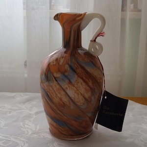 Könnte beinhalten: Eine braune und blaue Glasvase mit einem weißen Henkel. Die Vase hat ein Spiralmuster und steht auf einer weißen Tischdecke. Der Text "Die Kennkarte" befindet sich auf einem schwarzen Etikett.