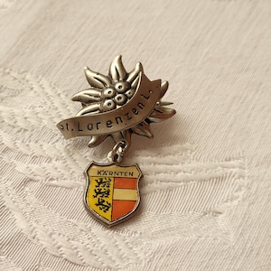 Könnte beinhalten: Silberne Anstecknadel mit einer stilisierten Edelweißblume und einem Banner mit der Aufschrift "St. Lorenzen L." Die Nadel ist an einem Schild mit dem Wappen von Kärnten, Österreich, befestigt. Das Schild ist gelb mit einem roten Streifen und drei gelben Löwen.