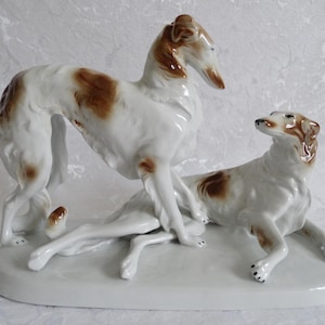 May include: A porcelain figurine of two white and brown hound dogs. The dogs are in a playful pose, with one dog standing on its hind legs and the other dog lying down. The figurine is on a white base.