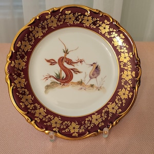 May include: A decorative plate with a burgundy rim and gold accents. The center of the plate features a hand-painted design of a seahorse and a bird.