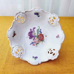 May include: A white porcelain bowl with a floral design and gold trim. The bowl has a scalloped edge and openwork details. The floral design features a bouquet of purple, red, and yellow flowers.
