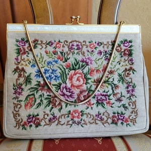 Puede incluir: Un bolso vintage beige con una correa de cadena dorada. El bolso está bordado con un patrón floral en tonos de rosa, morado, verde y azul.