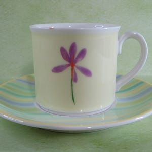 May include: A cream-colored ceramic coffee cup with a purple flower design and a matching saucer. The saucer has a striped pattern in shades of blue, green, and yellow. The cup has a white handle and rim.