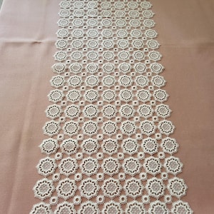 May include: White crocheted table runner with a delicate floral pattern. The runner is made of a fine, lightweight material and has a scalloped edge.