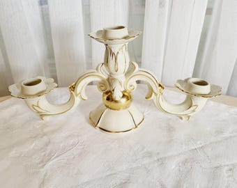 May include: Cream-colored, three-arm candelabra with gold accents. The ornate design features a central column with two curved arms, each holding a candle cup. The base and details are highlighted with gold trim, creating an elegant aesthetic.