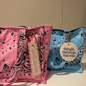 May include: Two tote bags, one pink and one blue, with a bandana print. The pink bag has a tag that says "cool mom." The blue bag has a tag that says "that girl. that mom. that wife."