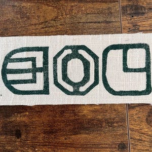 May include: A rectangular piece of off-white fabric with a dark green geometric design. The design consists of three abstract shapes: a rounded rectangle with horizontal lines, an octagon, and a square with a curved corner. The fabric has a raw edge.