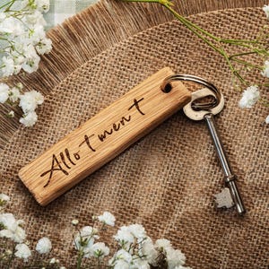 Könnte beinhalten: Ein rechteckiger Holzschlüsselanhänger mit dem eingravierten Wort Allotment in Schreibschrift. Der Schlüsselanhänger ist an einem silbernen Metallring befestigt, an dem ein einzelner silberner Schlüssel hängt. Das Holz hat eine sichtbare Maserung.