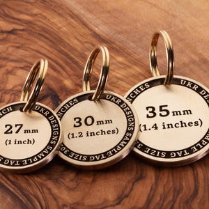 May include: Three brass keyrings with different sizes, 2.5 cm, 3 cm, and 3.5 cm. Each keyring has a round tag with the size printed in millimetres and inches. The text on the tag reads "UKR DESIGNS SAMPLE TAG SIZE".