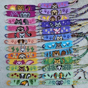 May include: A collection of colorful friendship bracelets, each featuring a unique animal design. The bracelets are made of woven threads in various colors, including pink, purple, and green. Animals include a lion, elephant, cat, raccoon, dog, sloth, axolotl, and panda.