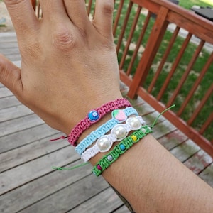 Pode incluir: Quatro pulseiras de amizade coloridas com contas e pingentes. As pulseiras são rosa, azul, branca e verde.
