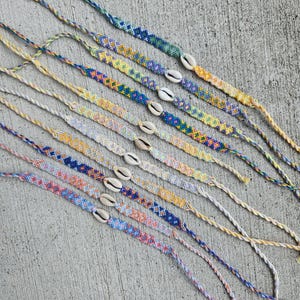 May include: A collection of colorful friendship bracelets with shell charms. The bracelets are made with a variety of colors, including blue, green, yellow, orange, and pink. The shells are a natural white color and are attached to the bracelets with a knot.