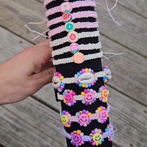 May include: A collection of colorful handmade bracelets displayed on a black cylindrical stand. The bracelets feature various designs, including fruit slices, smiley faces, and seashells. The bracelets are made with macrame knots and beads.