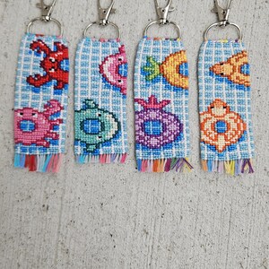 May include: Four handmade keychains with colorful embroidered sea creature designs. Each keychain features a different sea creature, including a red crab, pink fish, green turtle, purple starfish, and orange fish, on a blue and white checkered background.