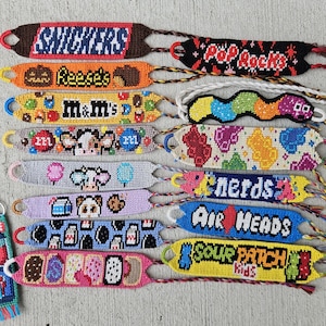 Puede incluir: Una colección de pulseras de amistad y llaveros hechos a mano y coloridos. Las pulseras presentan nombres de marcas de dulces y snacks como "Snickers", "Reese's" y "Air Heads". Los llaveros incluyen diseños de "Kit" e "Icee".